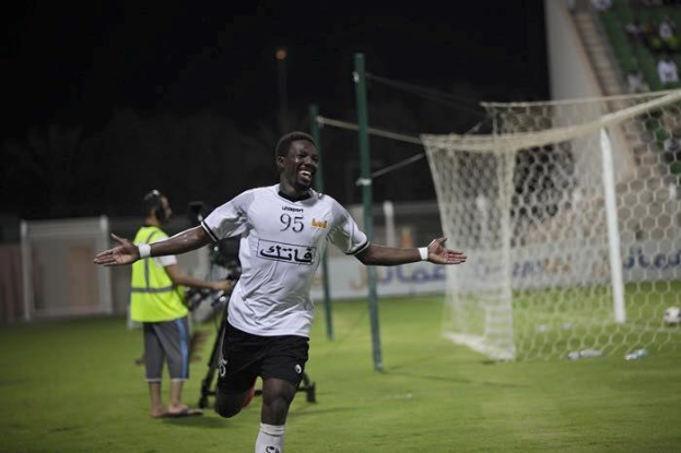 Ein Mann in weißem Hemd und schwarzen Shorts feiert auf einem Fußballfeld mit ausgestreckten Armen, umgeben von Zuschauern, mit einem Tor und einer Menge im Hintergrund.