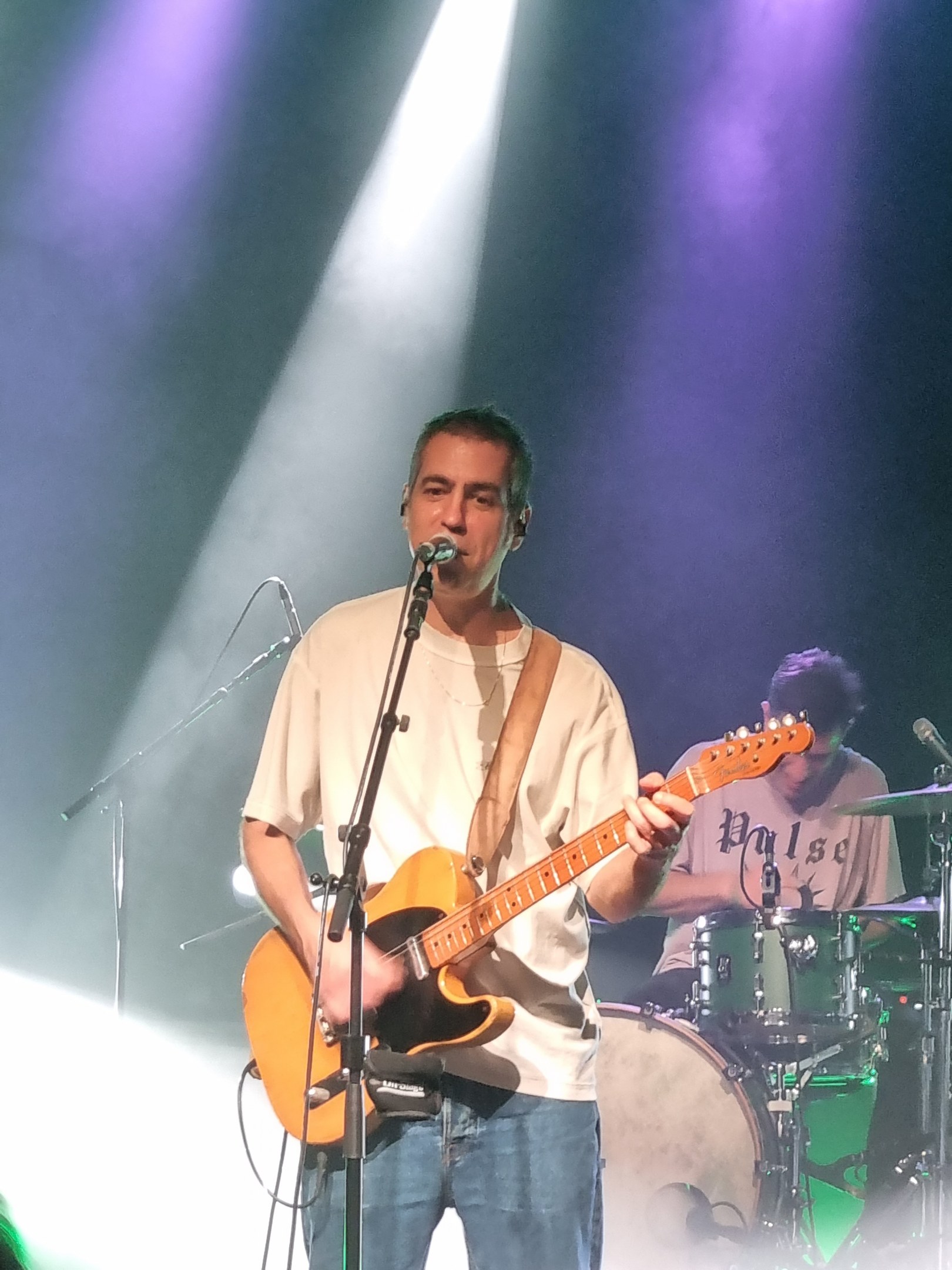 Ein Mann spielt eine E-Gitarre und singt in ein Mikrofon auf der Bühne, während ein anderer Musiker Schlagzeug spielt.