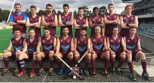 Eine Gruppe von Männern in Sportuniformen steht zusammen auf einem Feld und hält Hockeystöcke, feiert mit erhobenen Armen vor einem Stadion mit Zuschauern und einer Kulisse aus Bäumen unter einem klaren blauen Himmel.