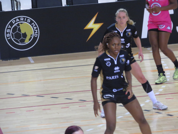 Gruppe von Frauen, die auf einem Platz Handball spielen, mit einem Banner, Stühlen und anderen Gegenständen im Hintergrund; eine Frau in Schuhen ist unten zu sehen.