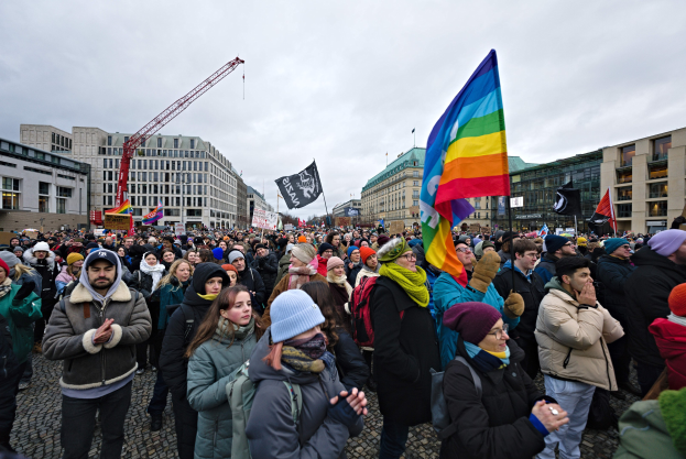 Eine große Gruppe von Menschen vor einem Gebäude mit Fahnen und Spruchbändern, darunter eines mit der Aufschrift "Lgbtq+ Rechte Demonstration in Berlin", einige tragen Mützen und Taschen, und Gebäude, ein Kran und Wolken im Hintergrund.