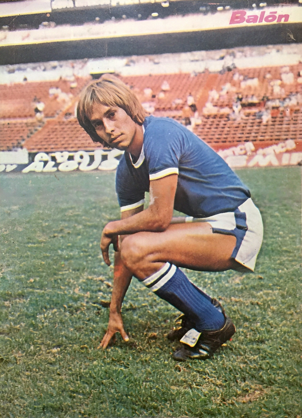 Ein Mann kniet auf einem Fußballfeld umgeben von Menschen auf Stühlen und Tafeln mit Text, trägt Schuhe.