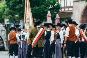 Eine Gruppe von Menschen in traditioneller bayerischer Tracht marschiert eine Straße entlang, einige halten Musikinstrumente und Fahnen, im Hintergrund sind Bäume, Gebäude und ein Auto zu sehen.