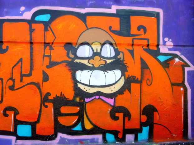 Graffiti-Gemälde eines Cartoon-Charakters mit großen Augen, einem breiten Grinsen und einem verschmitzten Ausdruck an der Seite eines Gebäudes, mit leuchtenden Farben und einer markanten Begleittext.