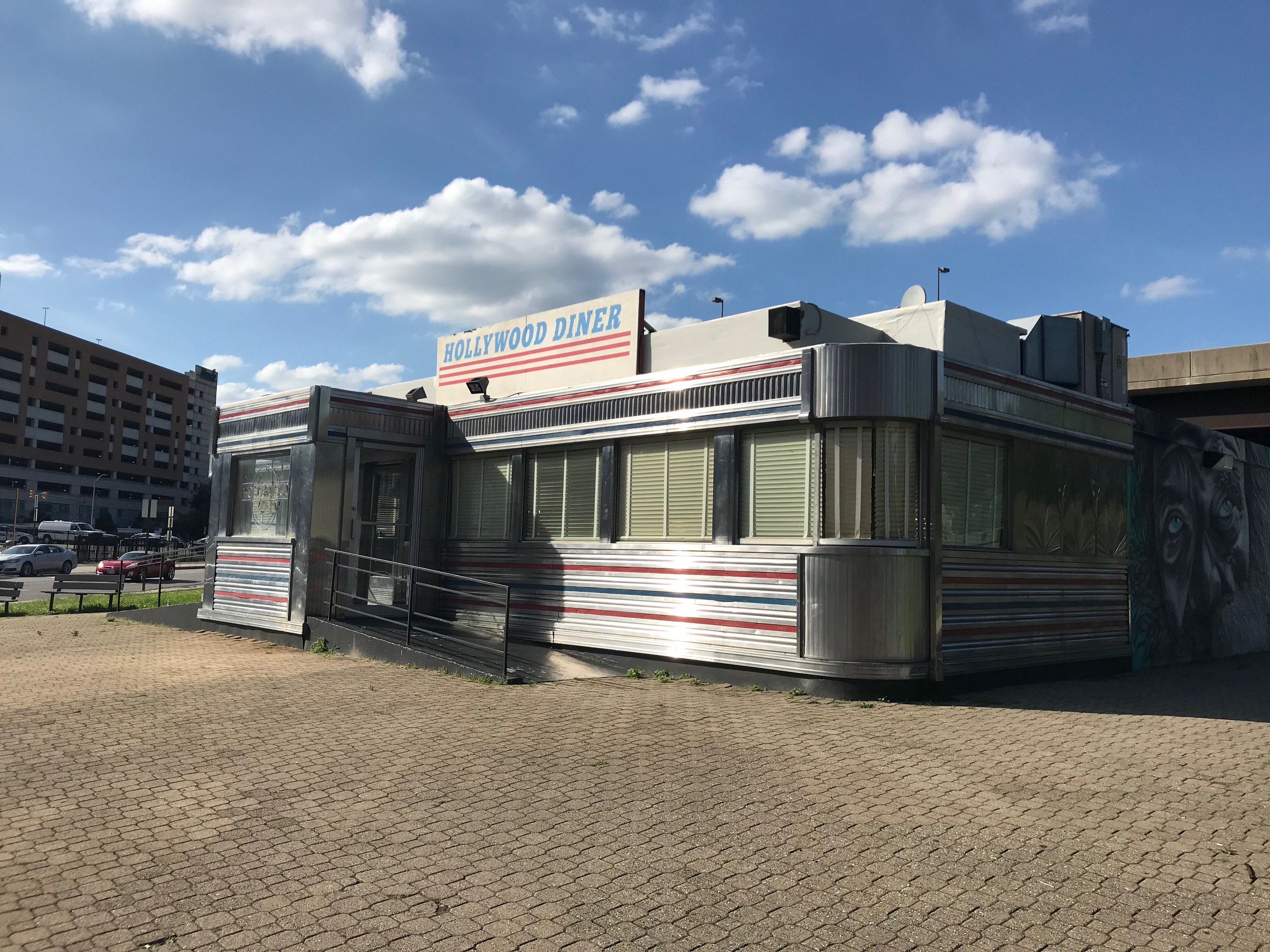 Außenansicht des Hollywood Diners mit seiner Leuchtschrift, umgeben von Gebäuden, Straßenmöbeln und Fahrzeugen unter einem bewölkten Himmel.