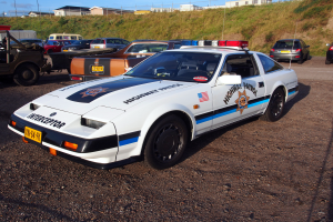Ein Polizeiwagen auf einem Parkplatz neben anderen Fahrzeugen, Gras, Pfählen, Drähten, Gebäuden und einem klaren blauen Himmel.