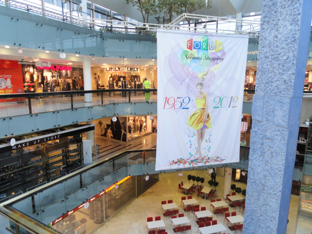 Großes Banner hängt von der Decke in einem Einkaufszentrum, mit zentral angeordneten Tischen und Stühlen, einer Säule auf der rechten Seite und einem Baum oben; Läden mit Schaufensterpuppen und ein paar Menschen sind im Hintergrund zu sehen.