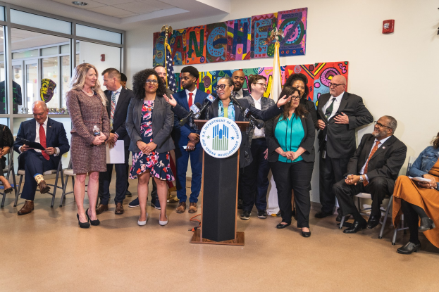 Gruppe von Menschen vor einem Podium mit einem Mikrofon und einer Tafel mit Text, einige sitzen auf Stühlen und andere stehen, mit Flaggen, einer Tafel, einem Feuerlöscher und Deckenlampen an der Wand.