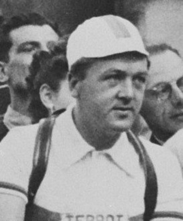 Schwarzes und weißes Foto eines Mannes in einer Mütze und Hosenträgern, der vor einer Gruppe von Menschen steht, die mutmaßlich ein legendärer Fußballspieler ist.