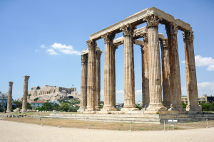 Alter Tempel von Olympie in Athen, Griechenland, mit seinen hohen korinthischen Säulen, umgeben von Bäumen und Felsen, mit einer Burg und bewölktem Himmel im Hintergrund.