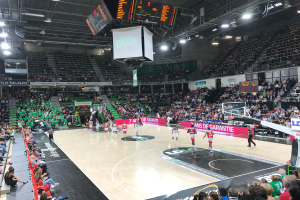 Ein Basketballspiel in einer großen Arena mit Zuschauern auf den Rängen, Anzeigetafeln mit "Asvel vs Galatasaray" und einer beleuchteten Decke mit einem Bildschirm über dem Platz.