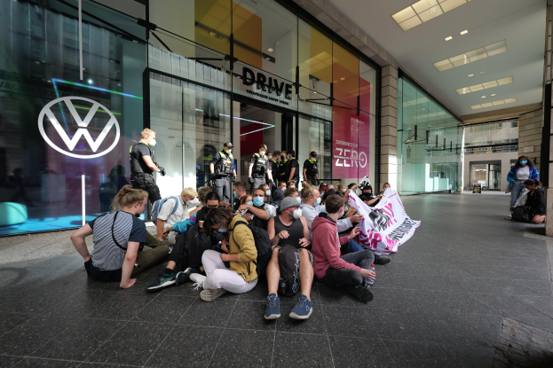 Eine Gruppe von Menschen sitzt vor einer Volkswagen-Vertretung mit Glaswänden auf dem Boden und hält ein Transparent mit Text, unter Deckenleuchten.