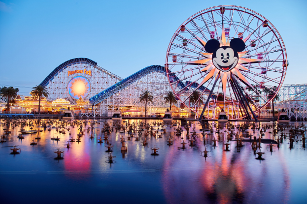 Eine farbenfrohe Szene im Disneyland California Adventure bei Sonnenuntergang mit einem riesigen Mickey-Maus-Rad im Vordergrund, einer Achterbahn im Hintergrund und Booten auf dem Wasser, mit einem bunten Himmel und Silhouetten von Bäumen.