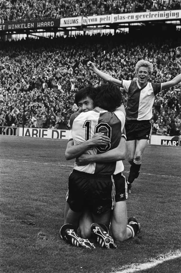 Drei Fußballspieler umarmen sich auf dem Boden und feiern ein Tor, mit einer jubelnden Menge und schwenkenden Fahnen im Hintergrund, dargestellt in Schwarz-Weiß.