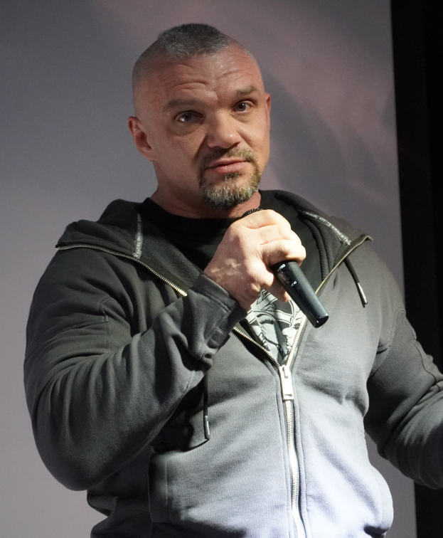 Ein Mann in schwarzer Kapuzenjacke und schwarzem Anzug mit weißem Hemd, der ein Mikrofon hält und selbstbewusst vor einer Wand steht.