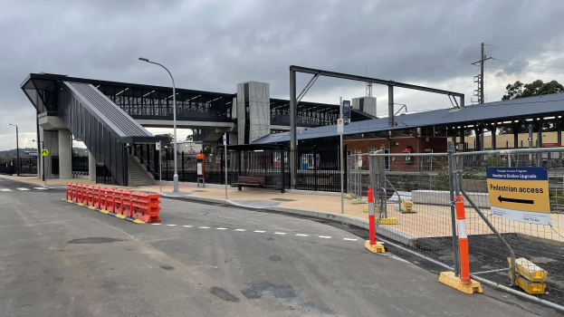 Ein belebter Bahnhof mit einer Brücke im Hintergrund, Straßeninfrastruktur, Gebäuden, Fahrzeugen und Bäumen unter einem bewölkten Himmel.